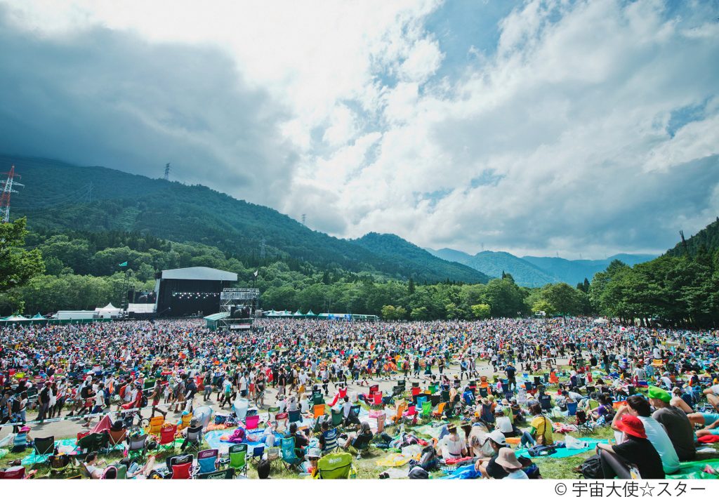 The landscape of mountains and trees wrap around this outdoor festival and crowds of people.