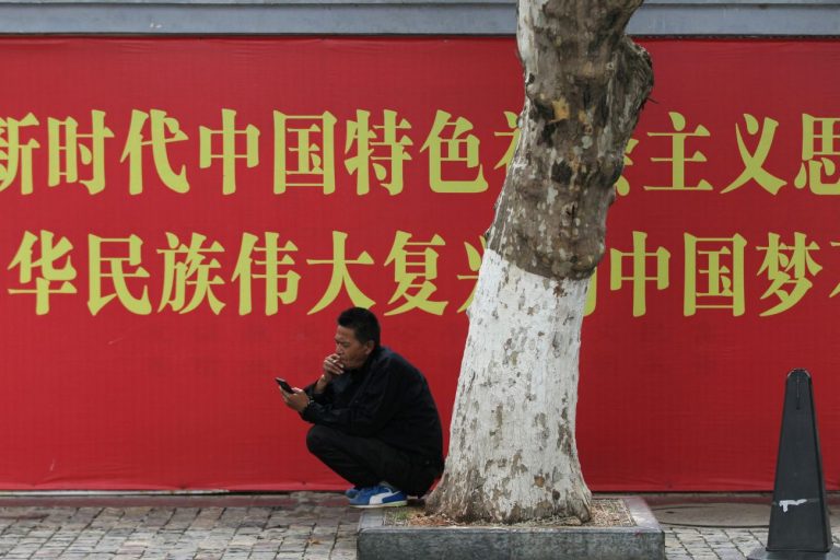 Man smokes a cigarette while looking at his mobile phone in front of a propaganda banner in Kunming